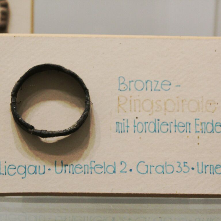 Ringspirale einer Ausgrabung bei Liegau. Ein kleines rundes Ausstellungsstück mit blauer Beschriftung in der Vitrine zur Archäologie.