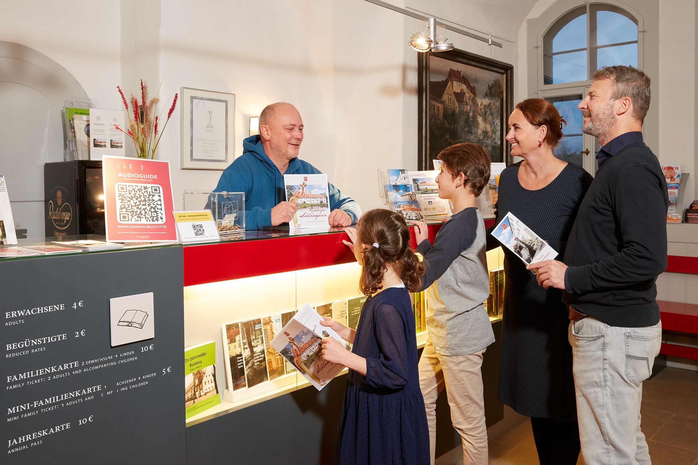 A family (parents and two schoolchildren) is standing at the counter of the castle cash desk in the entrance area of the castle.... A member of staff talks to them and shows them a booklet about the castle.