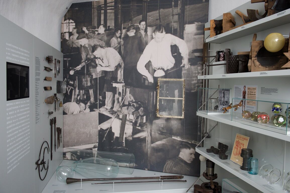 In der Mitte des Fotos sieht man ein wandhohes Foto, welches eine Arbeitssituation in einer Glashütte zeigt. Links und rechts sind Glasmacherwerkzeuge, Arbeitsformen und Produkte aussgestellt.