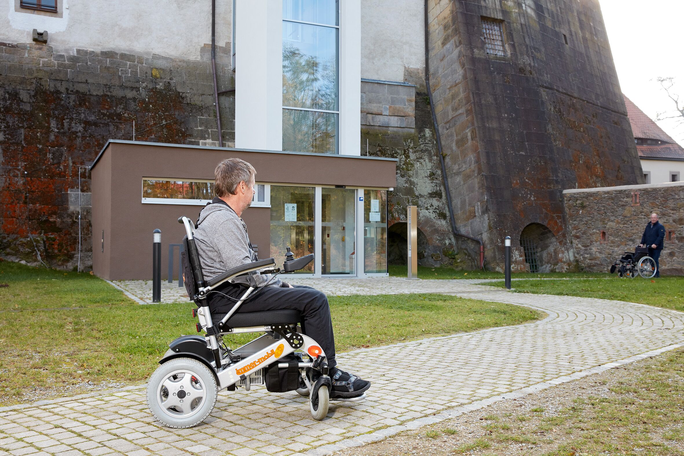 Ein Rollstuhlfahrer auf dem glatt gepflasterten Weg im Schlossgarten. Der Weg führt zum Aufzug.