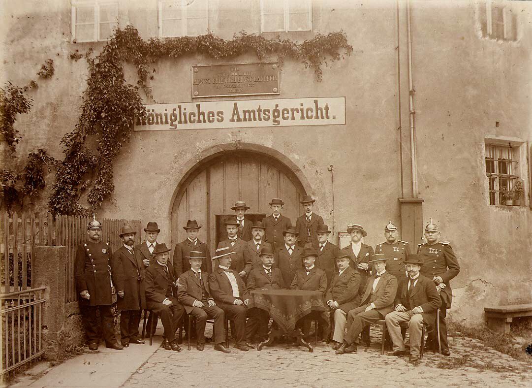 Foto in Vergrößerungsansicht öffnen: Auf einem Sepiafoto stehen und sitzen 32 Bedienstete des Amtsgerichtes vor dem Stadttor des Schlosses.