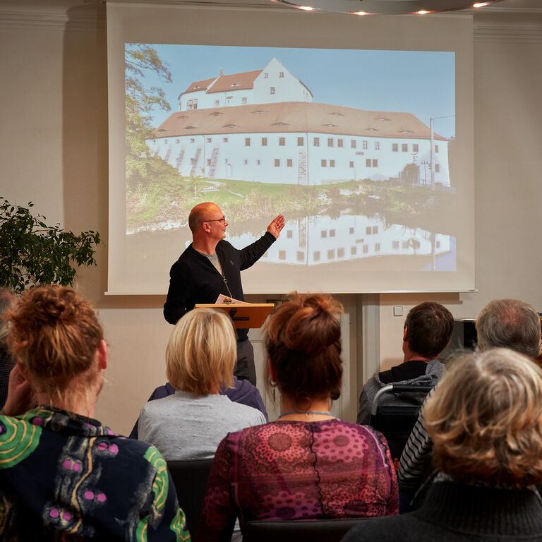 Der Sammlungsleiter hält im Festsal vor Publikum einen Vortrag über die Schlossgeschichte. Auf der Leinwand ist eine Schlossansicht zu sehen.