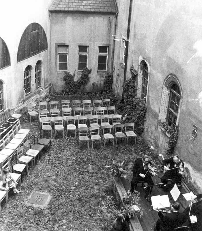 Innenhof mit mehreren leeren Stuhlreihen, einem kleinen Mädchen mit Puppe im Arm auf einem Stuhl sitzend  und einem kleinen Streichquartett auf einer erhöhten Bühne.