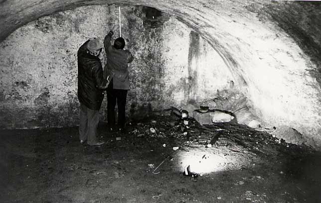 Das Schwarz-Weiß-Foto zeigt zwei Männer in Rückenansicht in einem Gewölbe. der Mann rechts misst die Deckenhöhe mit einem Zollstock. Am Boden liegt eine Taschenlampe.