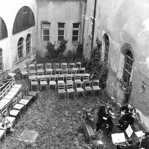 Innenhof mit mehreren leeren Stuhlreihen, einem kleinen Mädchen mit Puppe im Arm auf einem Stuhl sitzend  und einem kleinen Streichquartett auf einer erhöhten Bühne.