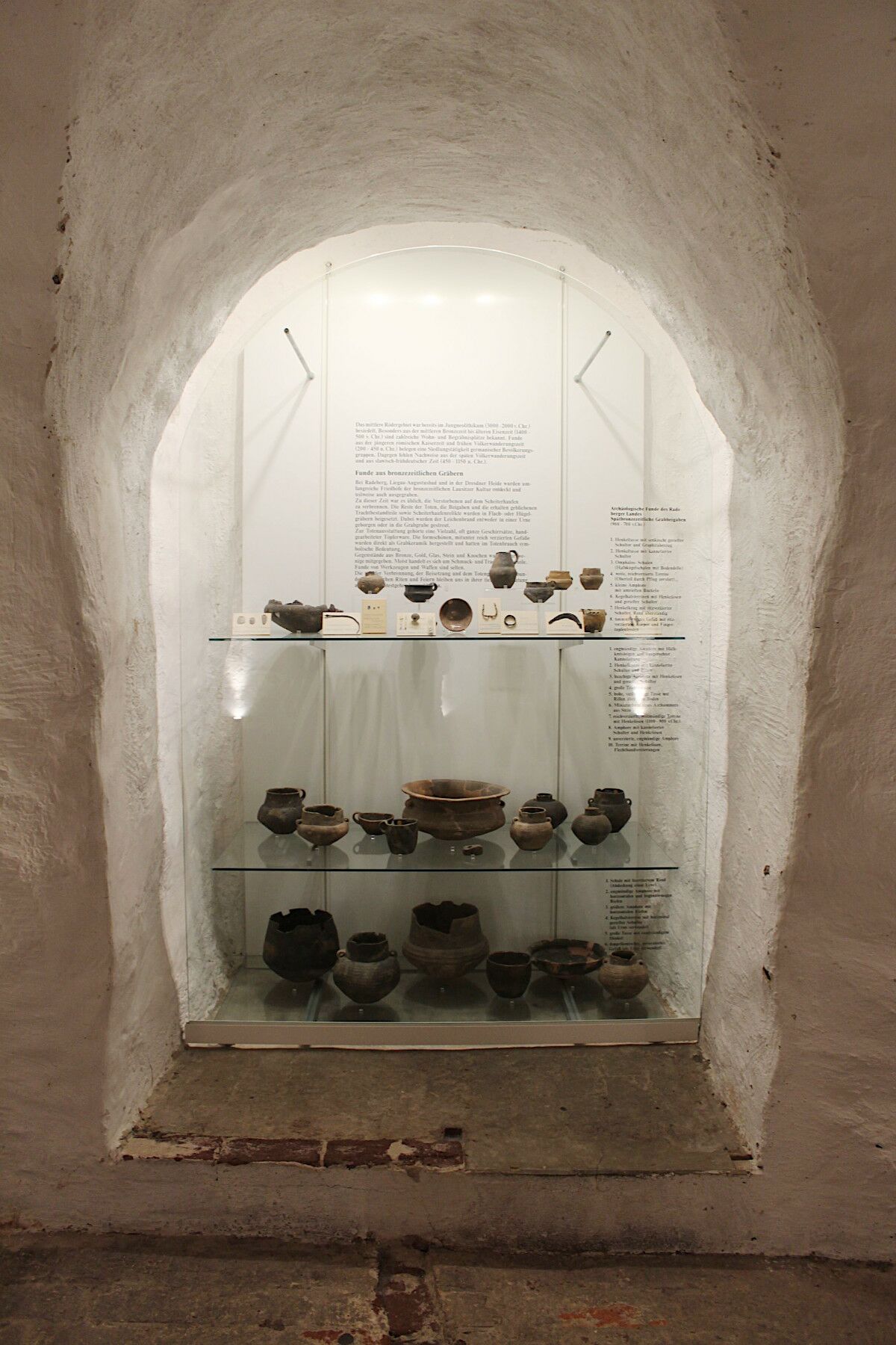 In einer hohen tiefen Wandnische befindet sich die Vitrine zum Thema Archäologie mit verschiedenen Fundstücken auf drei Ebenen..