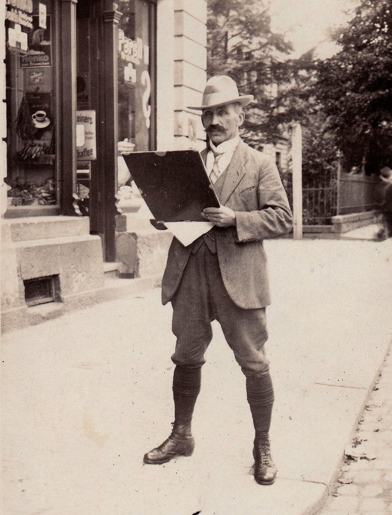Karl Stanka steht zeichnend auf einem Bürgersteig vor einem Geschäft. In der Hand hält er ein Zeichenbrett. Er trägt einen Anzug, Hut und Stiefel.