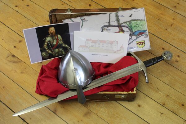 In einem geöffneteten braunen Koffer liegen auf einem roten Tuch ein Schwert und ein Ritterhelm, weiterhin ein Lageplan, eine Schlossansicht und ein Bildnis von Kurfürst Moritz.