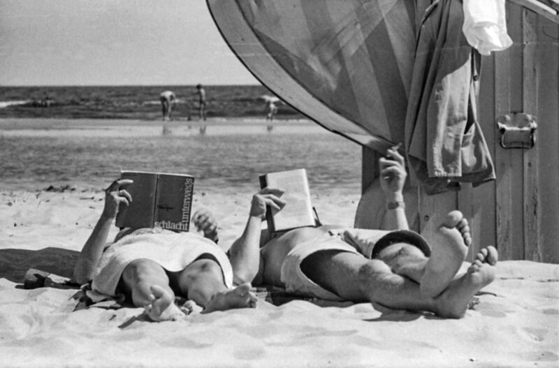 Zwei Menschen liegen auf dem Schwarz-weiß-Foto aus den 1960er Jahren am Strand. Sie liegen auf dem Rücken und halten in ihren Händen jeweils ein buch. Im Hintergrund ist das Meer zu sehen.