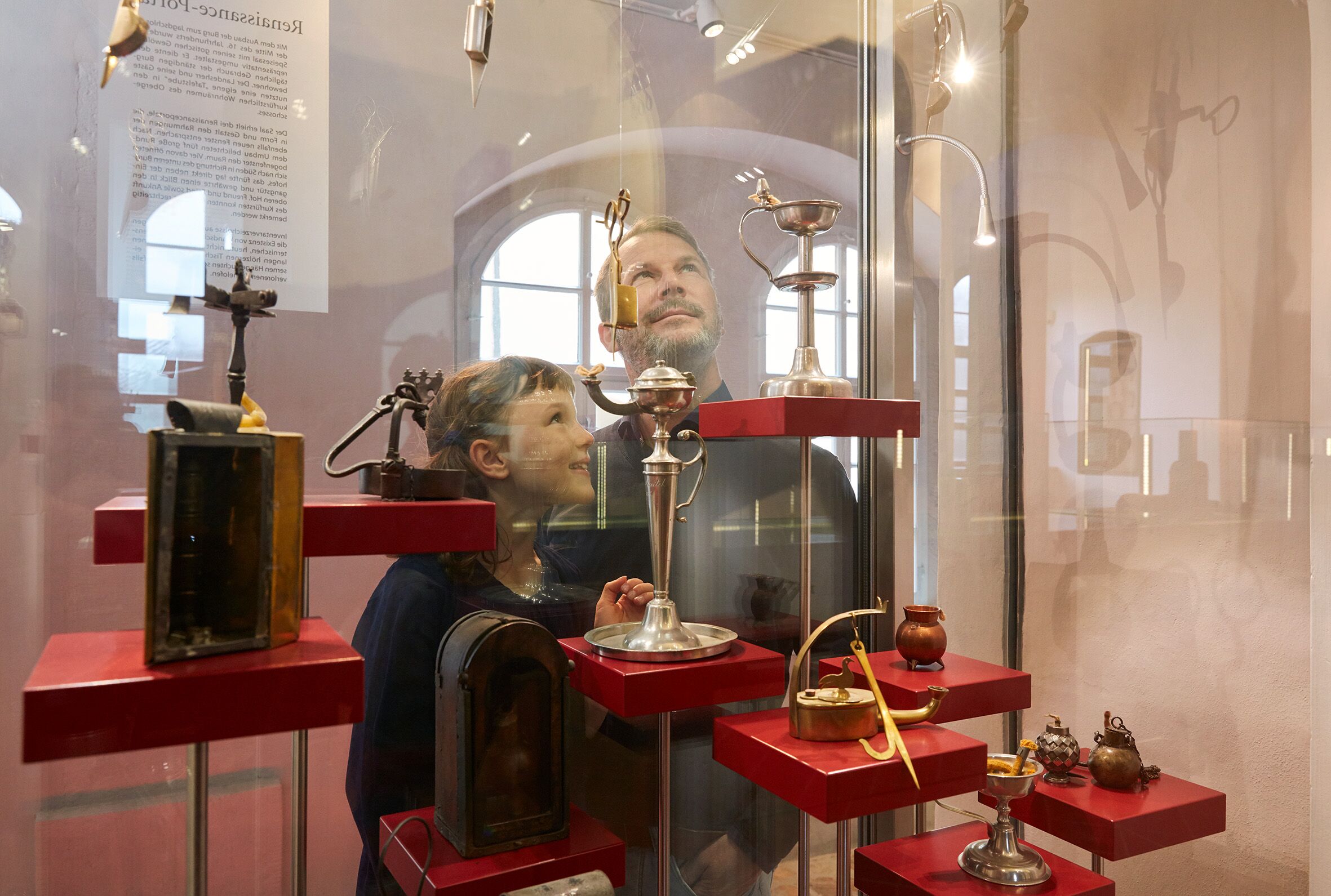 Foto in Vergrößerungsansicht öffnen: Hinter einer Glasvitrine mit Objekten aus edlen Metallen steht ein Mädchen mit ihrem Vater. Sie betrachten sich Exponate der Ausstellung.