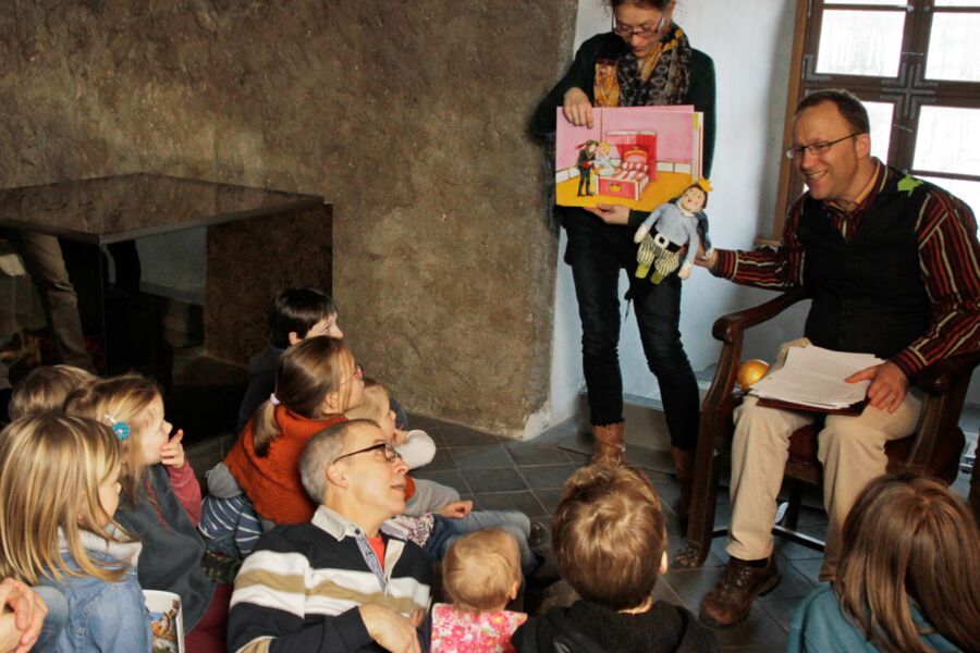Der Museumspädagoge liest einer Gruppe von Kindern das Märchen vom Froschkönig vor. Im Hintergrund zeigt eine Frau große Bildkarten, die das Märchen illustrieren.