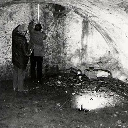 Das Schwarz-Weiß-Foto zeigt zwei Männer in Rückenansicht in einem Gewölbe. der Mann rechts misst die Deckenhöhe mit einem Zollstock. Am Boden liegt eine Taschenlampe.