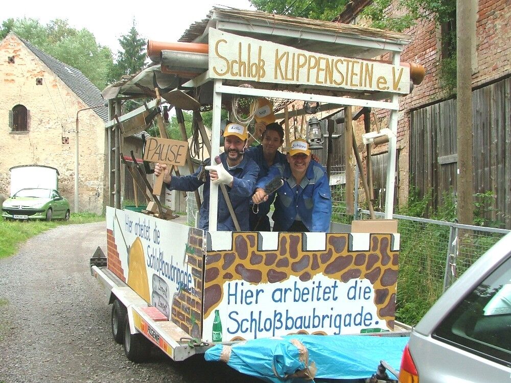 Drei Männer halten lachend auf dem selbstgebauten Umzugswagen ein Schild mit der Aufschrift Pause. Auf dem bunten Wagen steht geschrieben, hier arbeitet die Schlossbaubrigade.