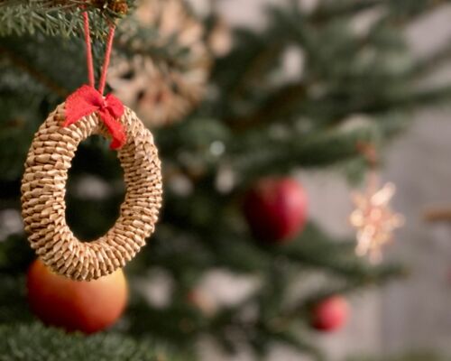 Geflochtener, runder Weihnachtsbaumschmuck mit rotem Band hängt an einem Tannenzweig, unscharfer Weihnachtsbaum im Hintergrund.