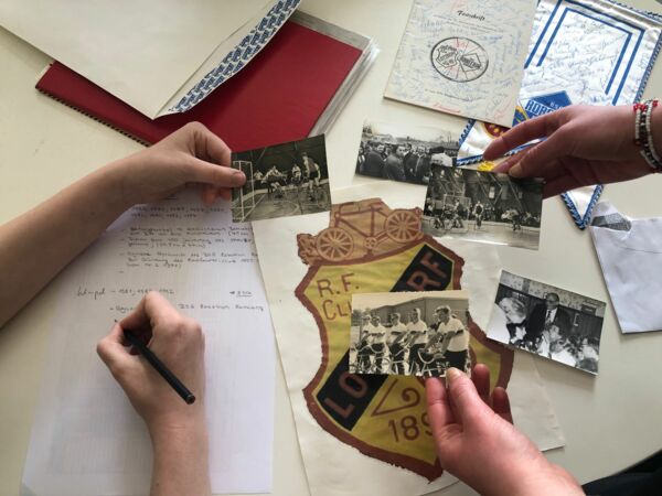 Vier Hände halten und ordnen alte Schwarz-Weiß-Fotos über einem Papier mit handschriftlichen Notizen und einem Wappen mit den Buchstaben 'R.F. Club' und dem Jahr 1895, Kontext 'genial sozial 2025'
