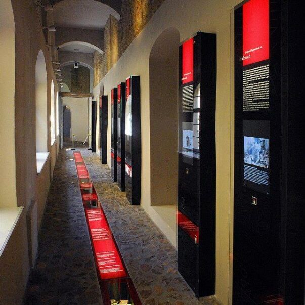 Blick in den Museumsgang. Am Boden befindet sich mittig ein roter Zeitstrahl. Rechts im Bild sind schwarze Wandstelen zu sehen mit Texten und Bildmonitoren.