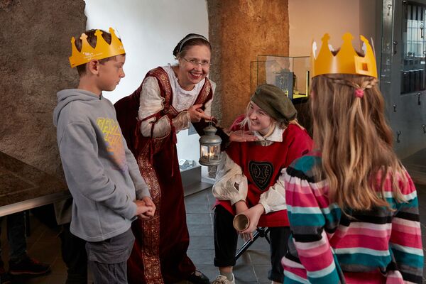 Unsere Museumspädagogin ist als Kurfürstin Agnes verkleidet und trägt ein langes, rotes Samtkleid. Sie erzählt über die Geschichte vom Schloss und von seinen Bewohnern. Drei Kinder lauschen gespannt.