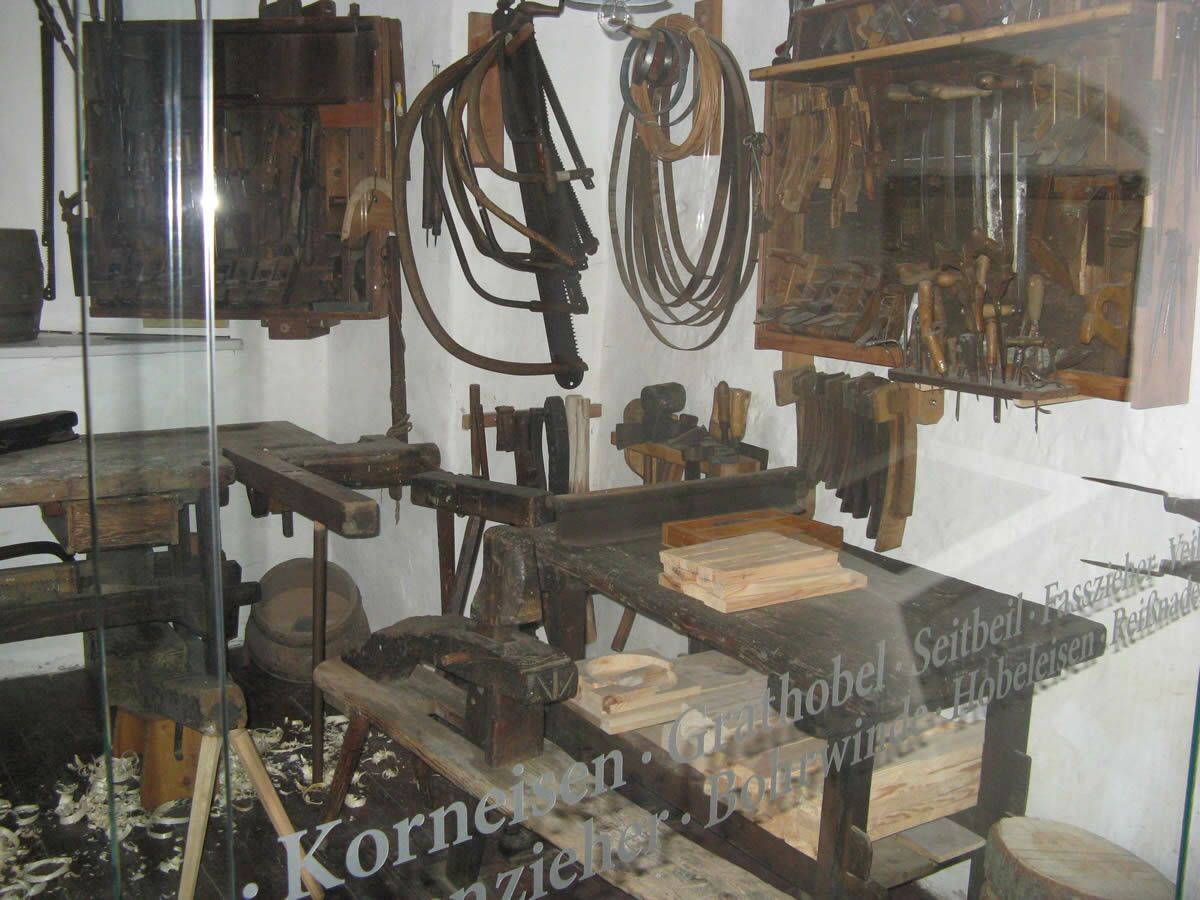 Blick in die Schauwerkstatt. Hinter einer großen Glasscheibe, die in der mitte zweizeilig mit Werkzeugbegriffen beschriftet ist, sieht man Werkbänke, Werkzeuge, Späne und Produkte aus Holz.