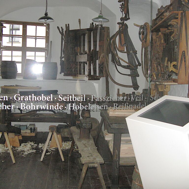 Blick durch die Glaswand in die Historische Böttcherei mit Arbeitsbänken und verschiedenen Werkzeugen auf Bänken und an der Wand.
