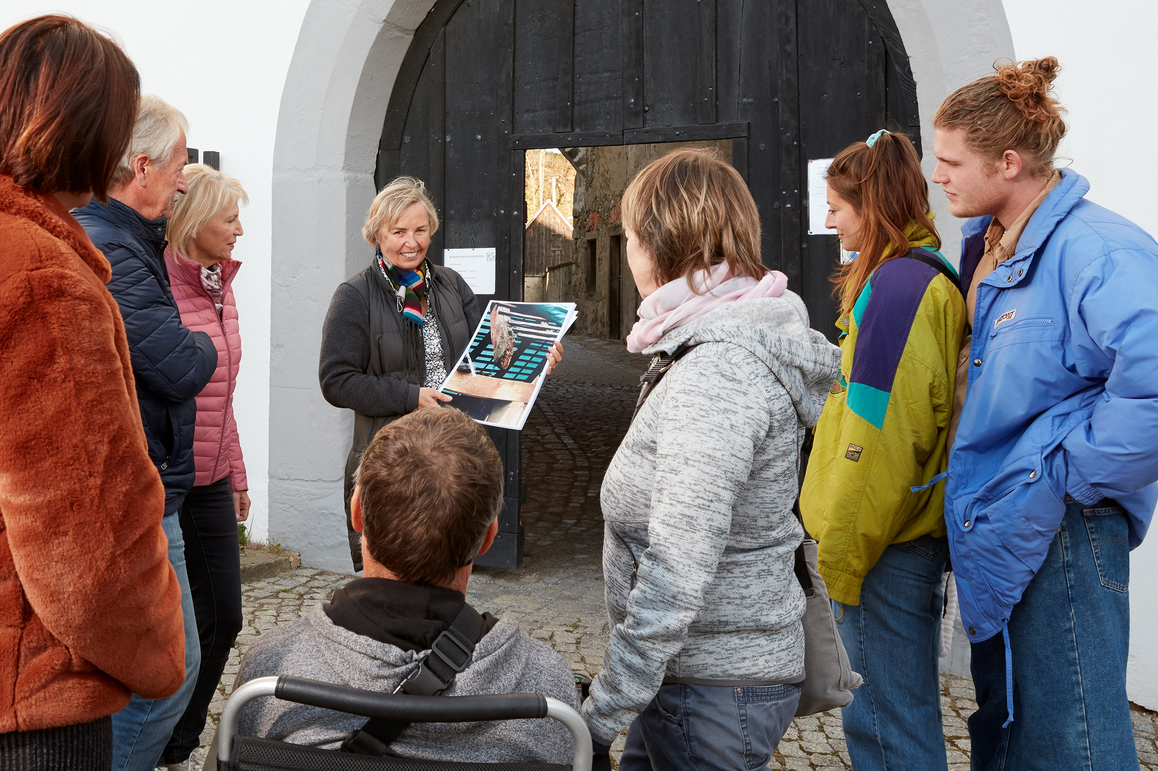 Die Schlossleiterin steht mit einer Gruppe von Besuchern vor dem großen Schlosstor. Durch die geöffnete Tür in der Mitte des schwarzen Tores kann man in den sonnigen Schlosshof sehen.