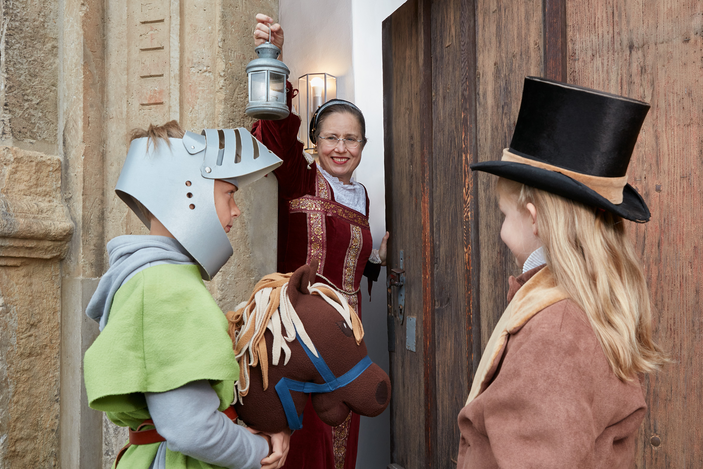 Die Museumspädagogin öffnet das hölzerne Tor mit erhobener Laterne. Sie trägt ein mittelalterliches Kostüm. Zwei verkleidete Kinder wollen hinein: Ein Junge mit Steckenpferd und Ritterhelm sowie ein Mädchen mit schwarzem Zylinder.