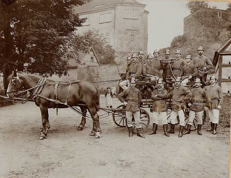 Feuerwehrleute von 1908 vor einem Wagen mit Wasserspritze, die von Pferden gezogen wird.
