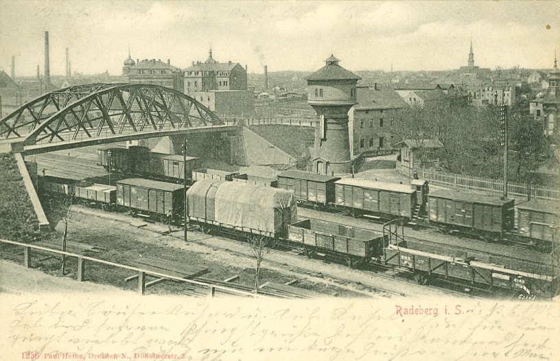 Foto in Vergrößerungsansicht öffnen: Die schwarzweiße Ansichtskarte zeigt einen Blick auf die Gleisanlagen in der Nähe des Radeberger Wasserturmes. Zwei Güterzüge fahren unter der Brücke durch. Im Hintergrund sieht man Industrieanlagen.