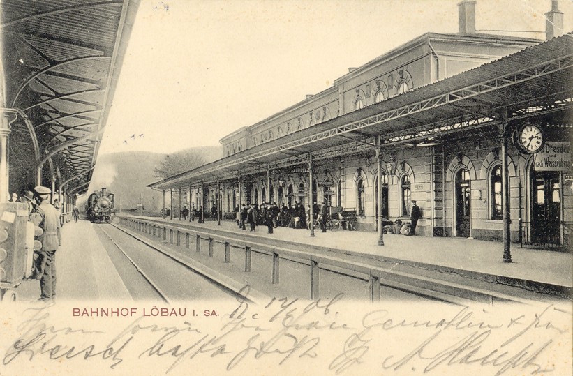 Foto in Vergrößerungsansicht öffnen: Ein Zug fährt in den Bahnhof Löbau ein. Blick vom Bahnhofsgebäude linke Seite über die beiden Gleise zum Bahnhofsgebäude rechte Seite. Dort stehen wartende Passagiere.