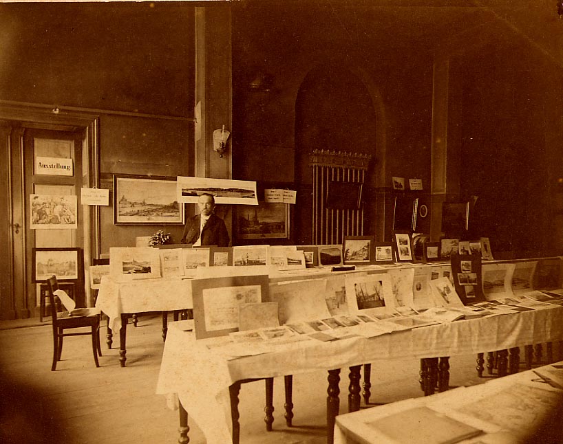 Eine Ausstellung in einem unbekanntn Saal um 1900. Auf drei Tafeln mit gedrechselten Beinen sind Grafiken ausgebreitet. Dahinter steht vor einer ebenfalls mit Bildern behangenen Wand ein Mann.